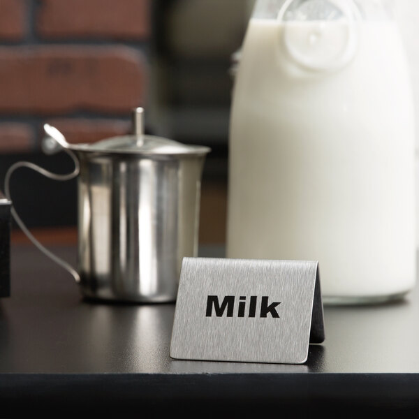 A Tablecraft stainless steel "Milk" tent sign in front of a metal pitcher of milk.