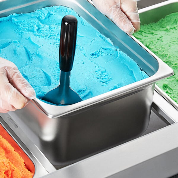 A person holding a stainless steel pan with blue and green ice cream using a metal scoop.