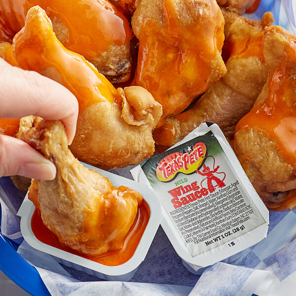 A hand dipping a Texas Pete Mild Wing Sauce cup into fried chicken.
