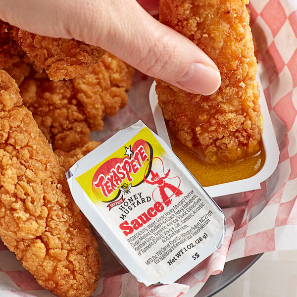 A person holding a Texas Pete Honey Mustard Dip Cup over fried chicken.