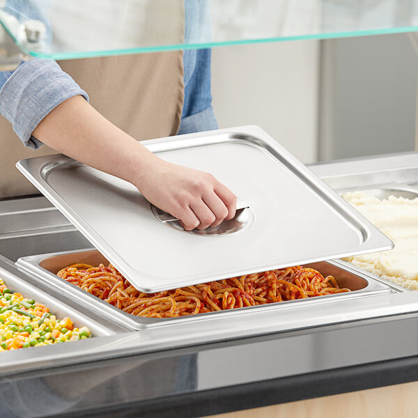 A person holding a Vigor 2/3 size stainless steel pan cover over a tray of food.