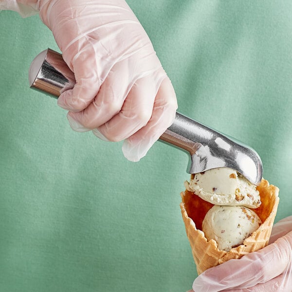 A hand in a glove using the Choice Aluminum Silver Ice Cream Scoop to add ice cream to a waffle cone.
