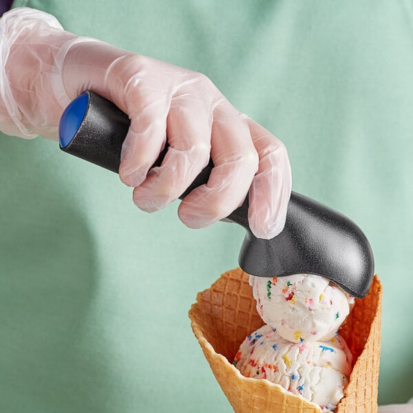 A person in a glove using a blue ice cream scoop to put ice cream in a waffle cone.