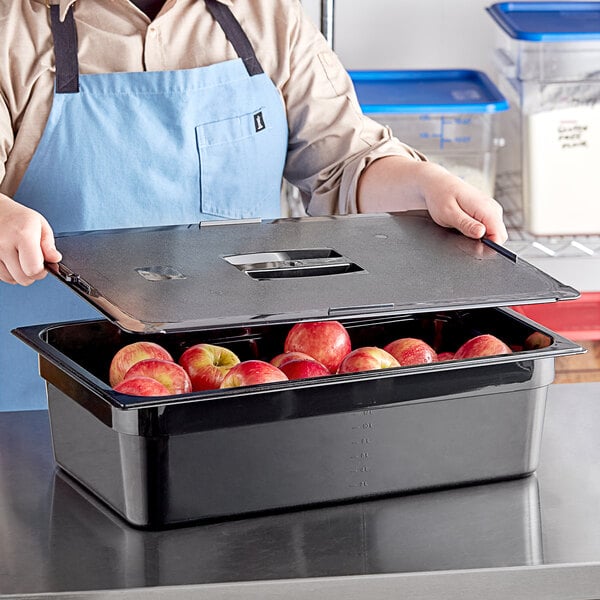 A person in a blue apron holding a black Vigor food pan with apples in it.