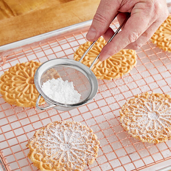 A person using a Choice stainless steel fine mesh strainer to sprinkle white powder over cookies.