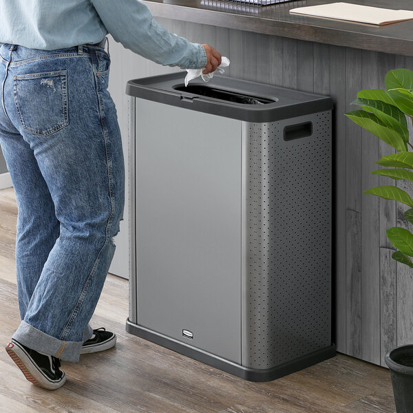 A person standing next to a Rubbermaid dark gray rectangular three-sided decorative trash can.