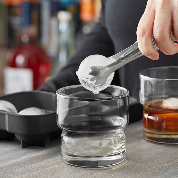 A person using tongs to place a clear sphere of ice in a glass.