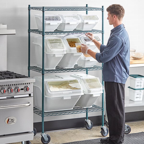 A man in a blue shirt and gloves using a white Baker's Mark shelving kit with clear bins to store food.