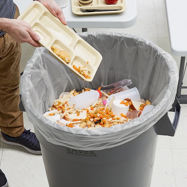 A person wearing a white shirt puts a tray of food into a trash can using a Lavex Hercules clear trash bag.