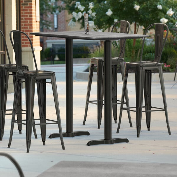 A Lancaster Table & Seating rectangular bar height table with two end outdoor base plates and black bar stools on a patio.