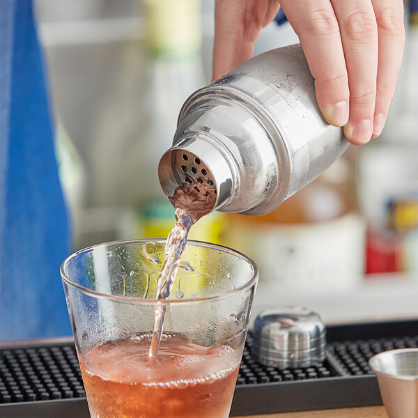 A hand pouring a Tablecraft stainless steel cocktail shaker into a glass of liquid.