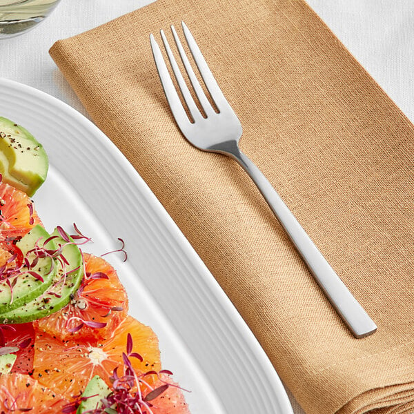 An Acopa Petra stainless steel fork on a napkin next to a plate of fruit salad.