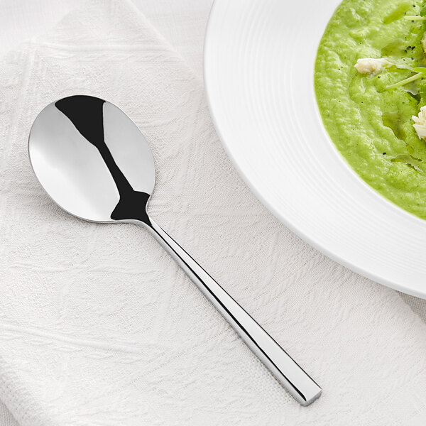 An Acopa stainless steel bouillon spoon next to a bowl of green soup on a white surface.