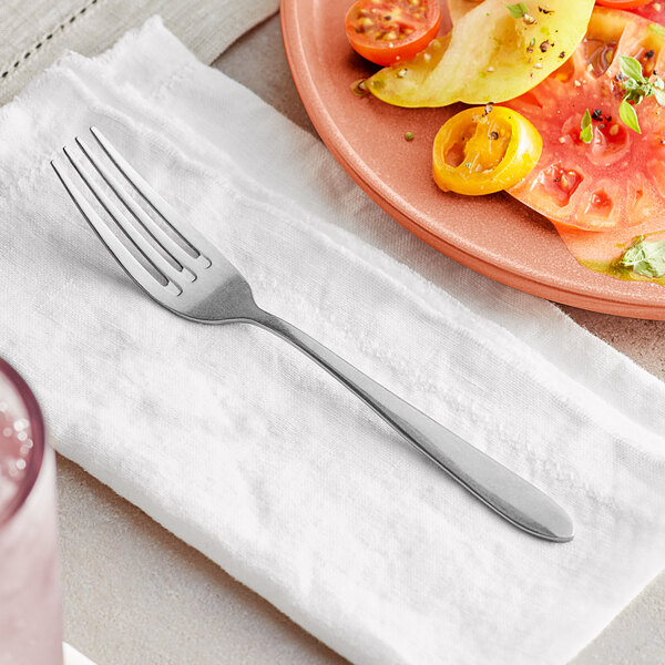 An Acopa Pangea stainless steel fork on a napkin next to a plate of tomatoes.
