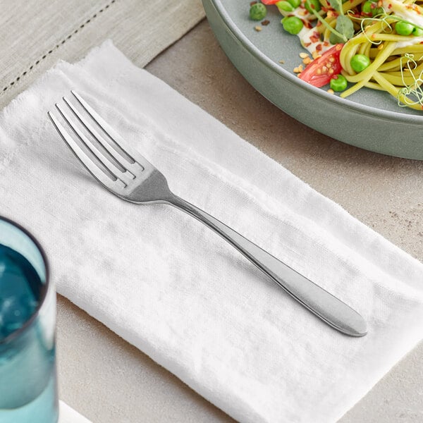 An Acopa Pangea stainless steel dinner fork on a white napkin next to a plate of food.