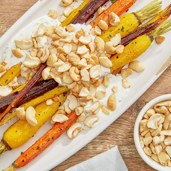 A plate of roasted carrots with cashew nuts and a bowl of nuts on a table.