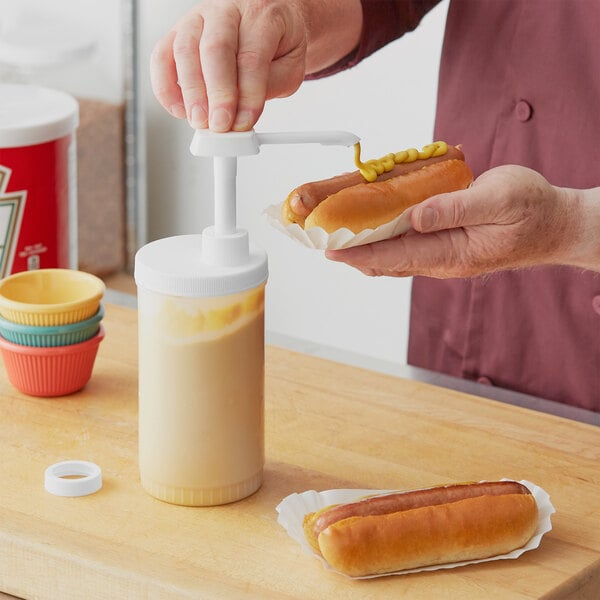 A person using a Choice red and white condiment pump to pour mustard onto a hot dog.