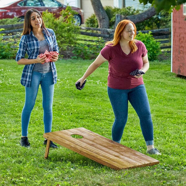 Two women playing a game of Backyard Pro rustic dark wood cornhole on grass.