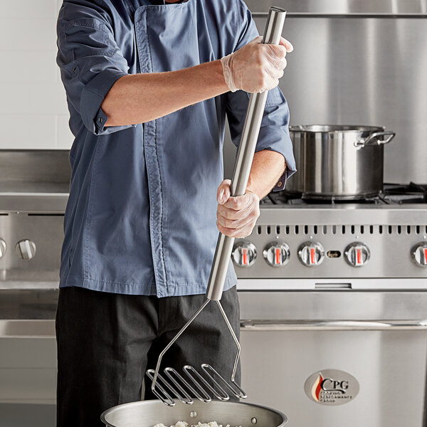 A man in a chef's uniform using a Tablecraft stainless steel potato masher to stir a pot of food.