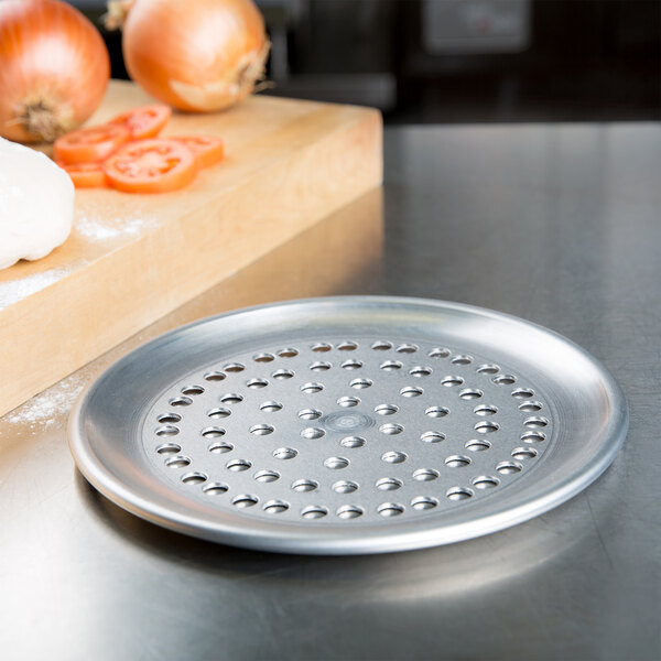 An American Metalcraft heavy weight aluminum pizza pan with perforations on a table next to onions.