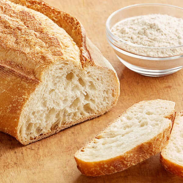 A bowl of white powder next to a loaf of bread on a cutting board.