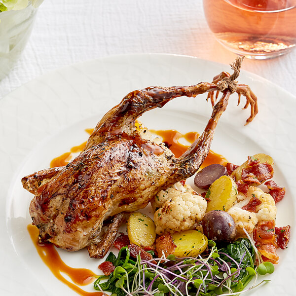 A white plate topped with a Manchester Farms fresh whole quail with vegetables and a glass of wine.