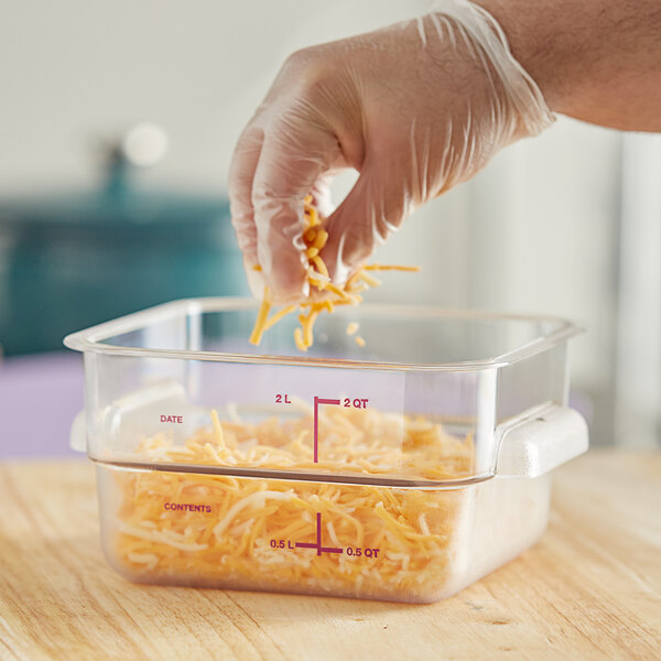 A person in gloves pouring shredded cheese into a Vigor clear polycarbonate food storage container.