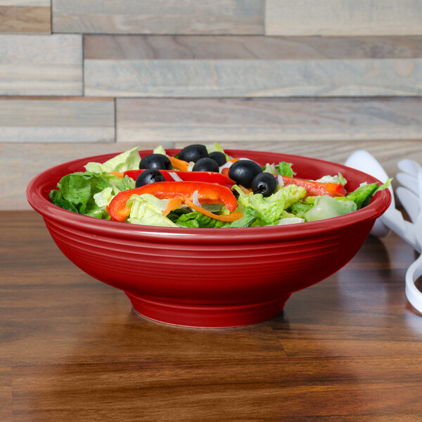 A close-up of a red Fiesta pedestal bowl filled with salad on a table.