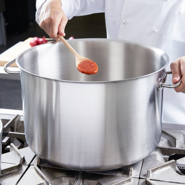 A person stirring red sauce with a wooden spoon in a large silver Vollrath sauce pot.