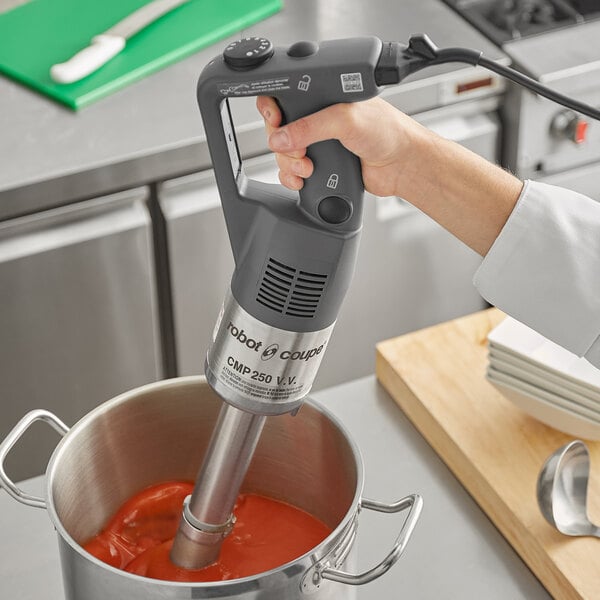 A hand using a Robot Coupe immersion blender to mix red sauce in a pot.