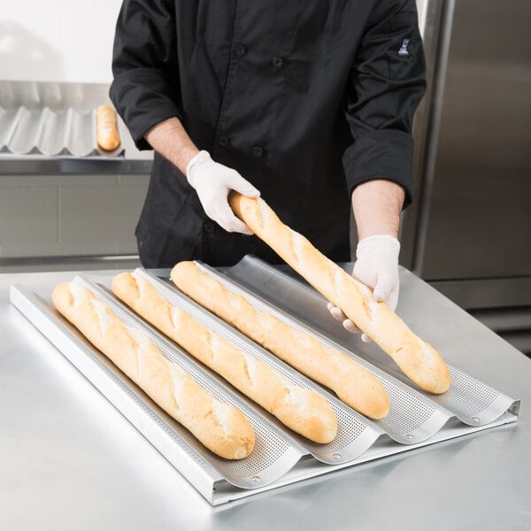 A man in a chef's uniform holding a tray of long French bread loaves.