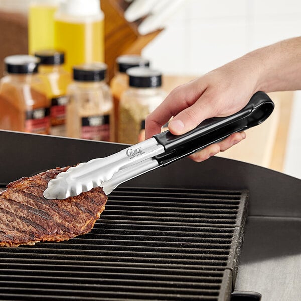 Choice black stainless steel scalloped tongs being used to cook meat on a grill.