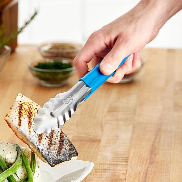 A person holding a blue Choice stainless steel scalloped tong over a grilling fish.
