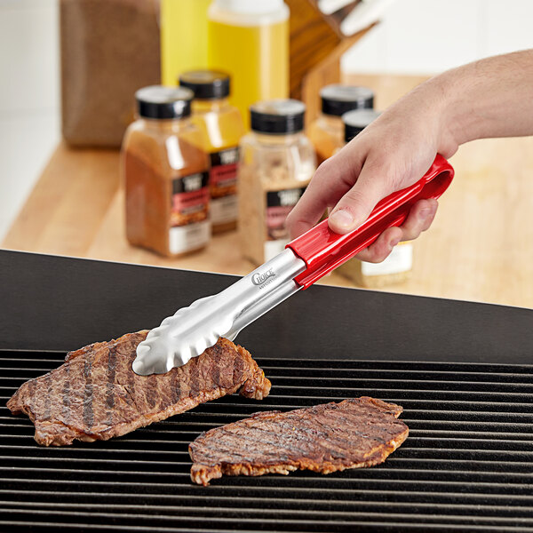 A hand holding Choice red stainless steel scalloped tongs over a grill.