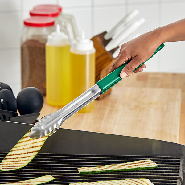 A hand using green and silver Choice scalloped tongs to cook zucchini on a grill.