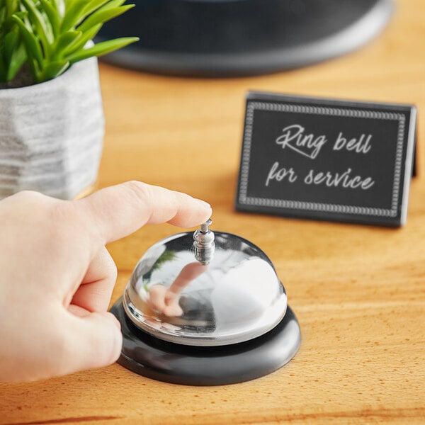 A hand ringing a Choice stainless steel call bell on a table.