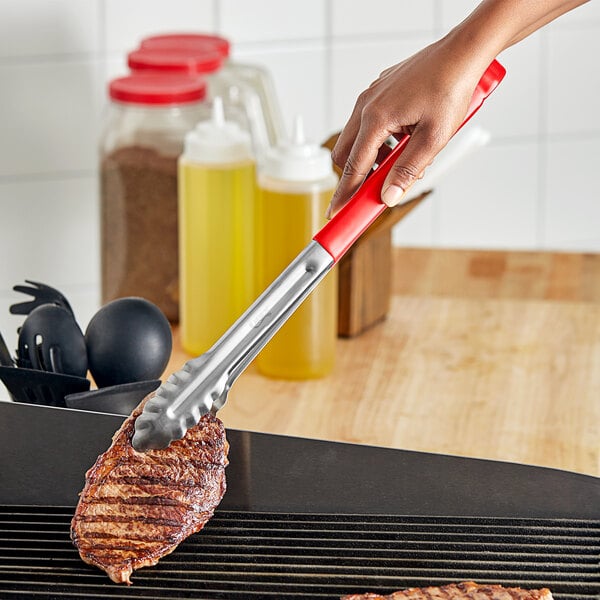 A hand using Choice red stainless steel scalloped tongs to grill meat.