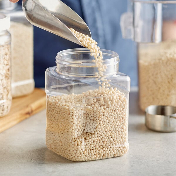 A person using a metal scoop to pour white grains into a square plastic jar.