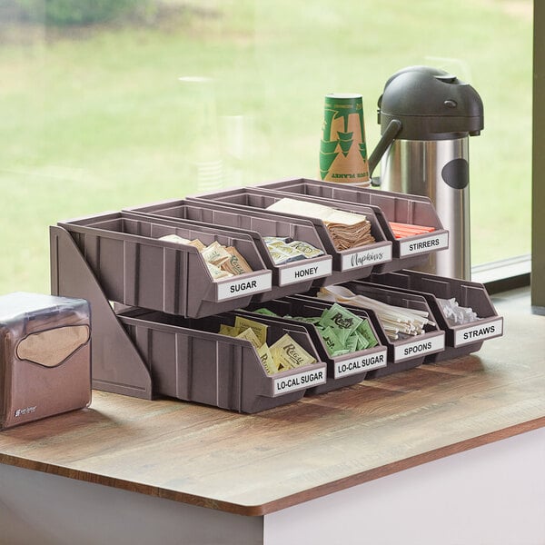 A brown 2-tier self-serve organizer set with bins on a counter with coffee cups and sugar.