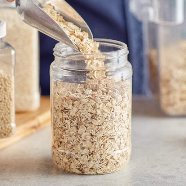 A person using a metal scoop to pour oats into a 32 oz. round PET plastic jar.
