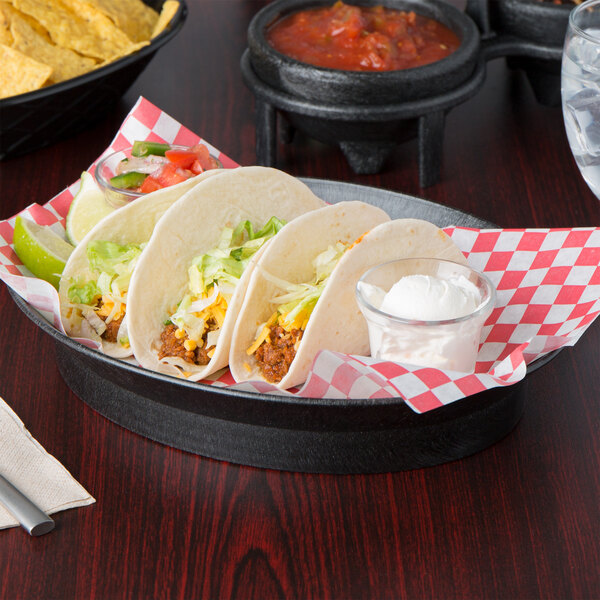 An oval bowl of tacos on a table with a bowl of chips and a red and white checkered napkin.