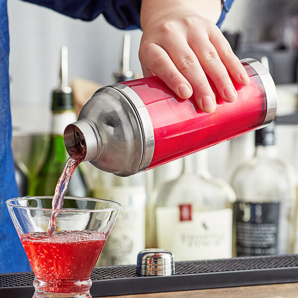A hand using a red and silver Franmara double wall cocktail shaker to pour a red drink into a glass.