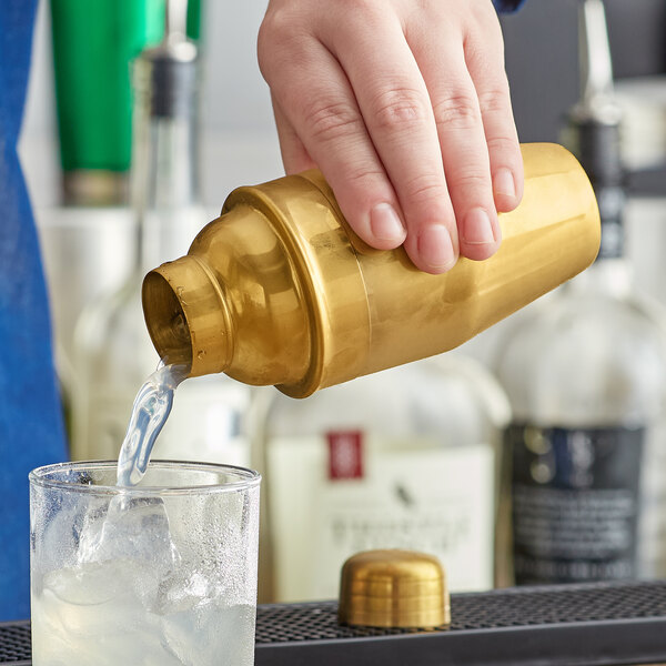 A hand using a Franmara gold-plated cocktail shaker to pour a drink into a glass.