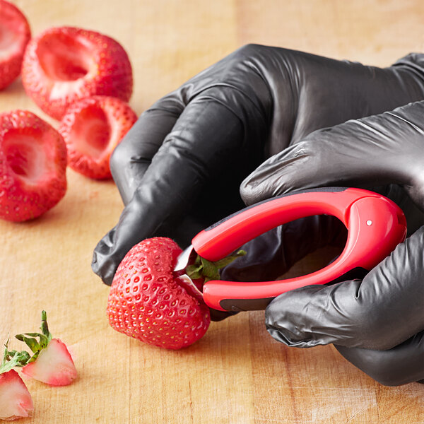 A pair of black gloves using an OXO Good Grips stainless steel strawberry huller to cut a strawberry.