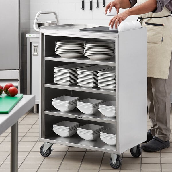 A man standing in a kitchen next to a Regency stainless steel utility cart with plates and bowls on the shelves.
