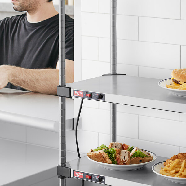 A man in a black shirt sits at a table with a Metro stainless steel heated shelf holding plates of a cheeseburger, fries, and a sandwich.