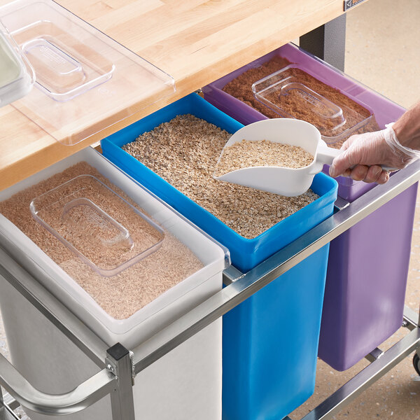 A person using a scoop to put food into a white, blue, and purple Baker's Mark ingredient bin.