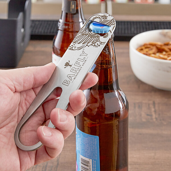 A person using a Barfly stainless steel speed bottle opener to open a brown bottle of liquid on a bar counter.