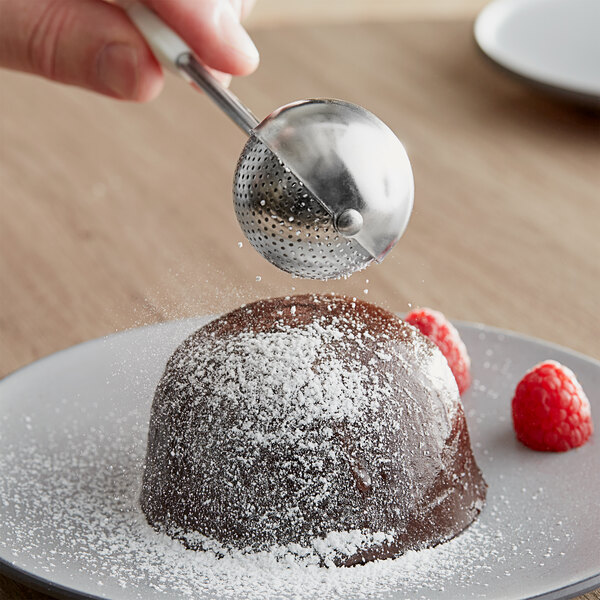 A hand holding an OXO Good Grips white and stainless steel dusting wand dusting powdered sugar on a chocolate dessert.