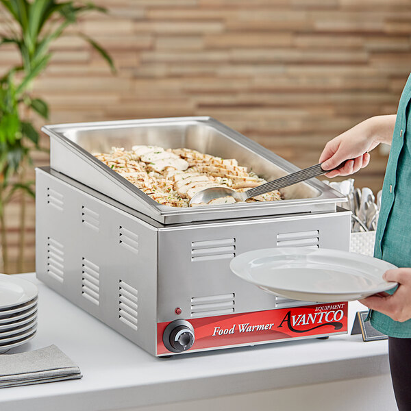 A woman holding a plate of food in front of an Avantco countertop food warmer.